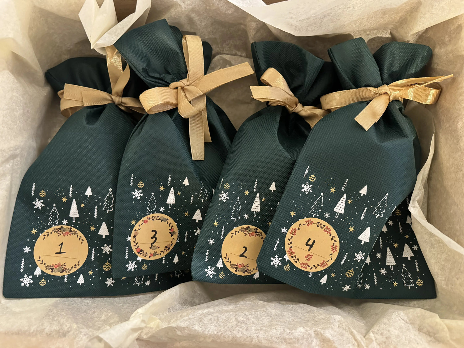 Komiko adventskalender 2025 – fire grønne stofposer nr. 1–4 med guldsløjfer i gaveæske med silkepapir.