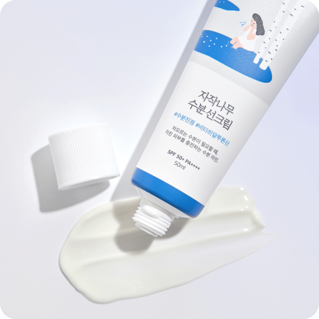 Round Birch Juice Moisturizing Sun Cream på hvid baggrund. En tube med fugtgivende solcreme, der er blevet åbnet, og noget af cremen er presset ud ved siden af tuben. Tuben har en blå og hvid etiket med koreansk skrift og en illustration af en person ved birketræer. SPF 50+ PA++++.