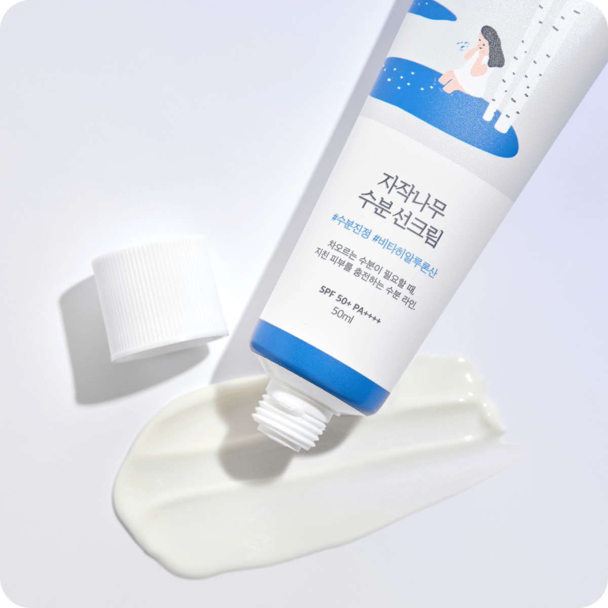 Round Birch Juice Moisturizing Sun Cream på hvid baggrund. En tube med fugtgivende solcreme, der er blevet åbnet, og noget af cremen er presset ud ved siden af tuben. Tuben har en blå og hvid etiket med koreansk skrift og en illustration af en person ved birketræer. SPF 50+ PA++++.