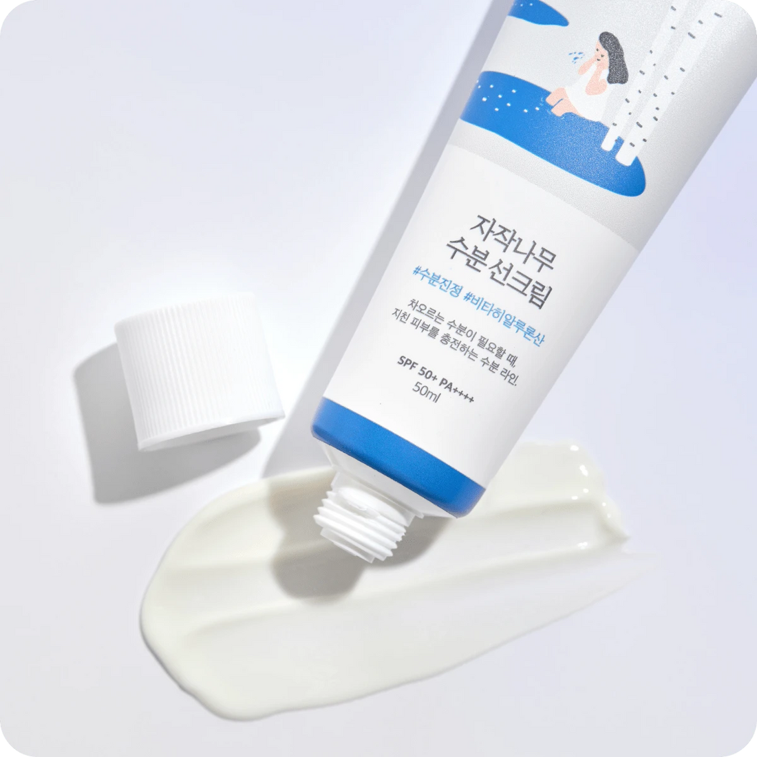 Round Birch Juice Moisturizing Sun Cream på hvid baggrund. En tube med fugtgivende solcreme, der er blevet åbnet, og noget af cremen er presset ud ved siden af tuben. Tuben har en blå og hvid etiket med koreansk skrift og en illustration af en person ved birketræer. SPF 50+ PA++++.